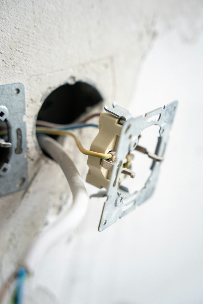 An exposed electrical wall socket in mid-installation, with visible wiring and mounting hardware set against a raw plaster wall. The image captures a hands-on moment in home renovation or electrical work.