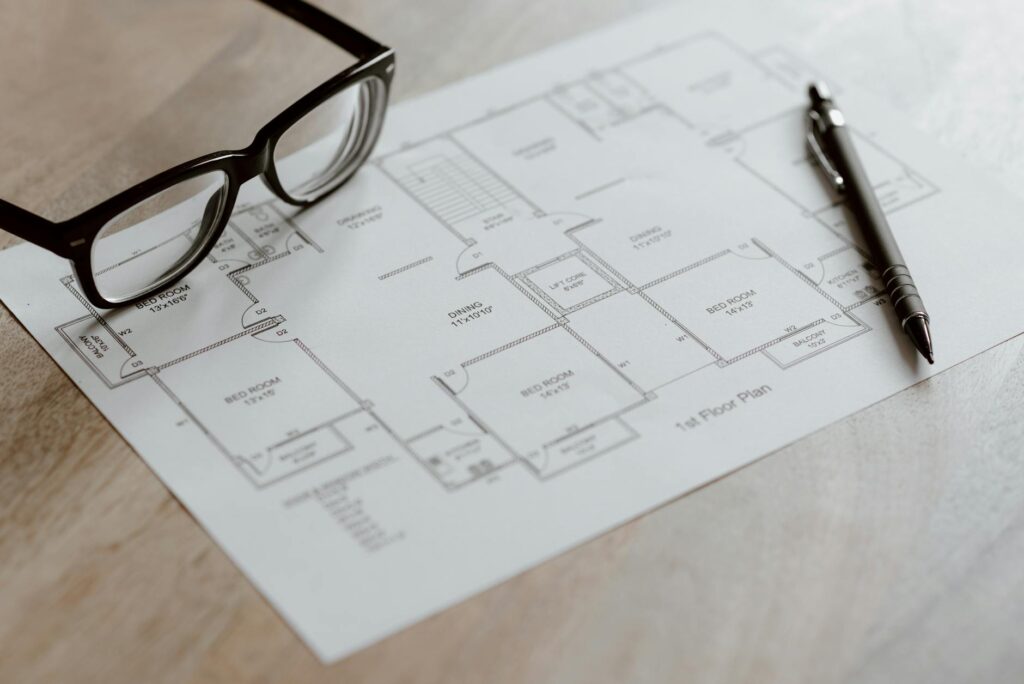 Composition of plan of room and black pen with eyeglasses placed on wooden table at daytime